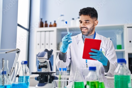 Young hispanic man wearing scientist uniform using touchpad holding test tube at laboratory