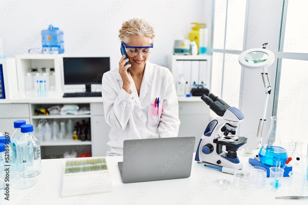 Middle age blonde woman wearing scientist uniform talking on the smartphone working at laboratory Middle age blonde woman wearing scientist uniform talking on the smartphone working at laboratory
