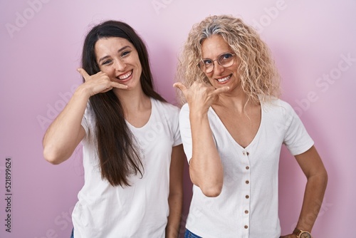 Mother and daughter standing together over pink background smiling doing phone gesture with hand and fingers like talking on the telephone. communicating concepts.