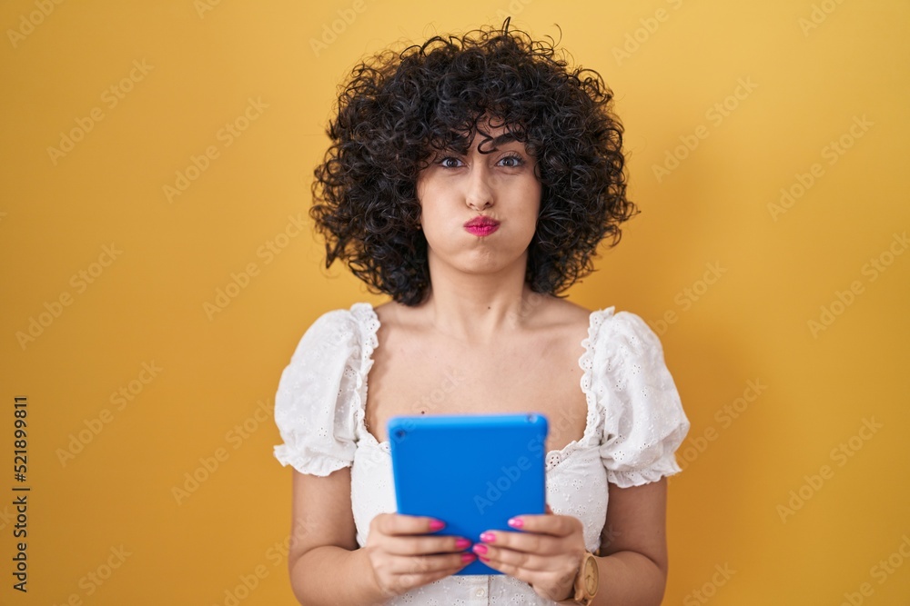 Young brunette woman with curly hair using touchpad over yellow background puffing cheeks with funny face. mouth inflated with air, catching air. Young brunette woman with curly hair using touchpad over yellow background puffing cheeks with funny face. mouth inflated with air, catching air.
