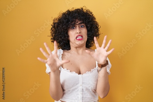 Young brunette woman with curly hair standing over yellow background afraid and terrified with fear expression stop gesture with hands, shouting in shock. panic concept.