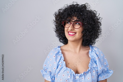 Young brunette woman with curly hair wearing glasses over isolated background smiling looking to the side and staring away thinking.