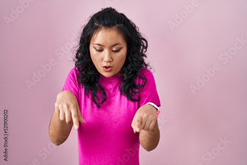 Young asian woman standing over pink background pointing down with fingers showing advertisement, surprised face and open mouth