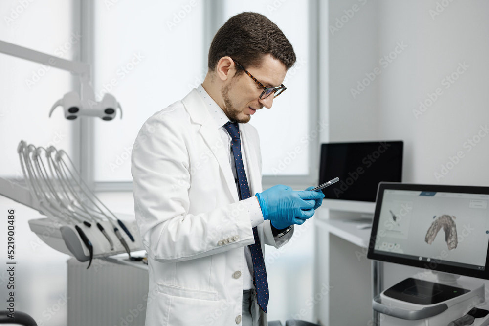 Fototapeta premium Handsome male dentist using smartphone in dental office