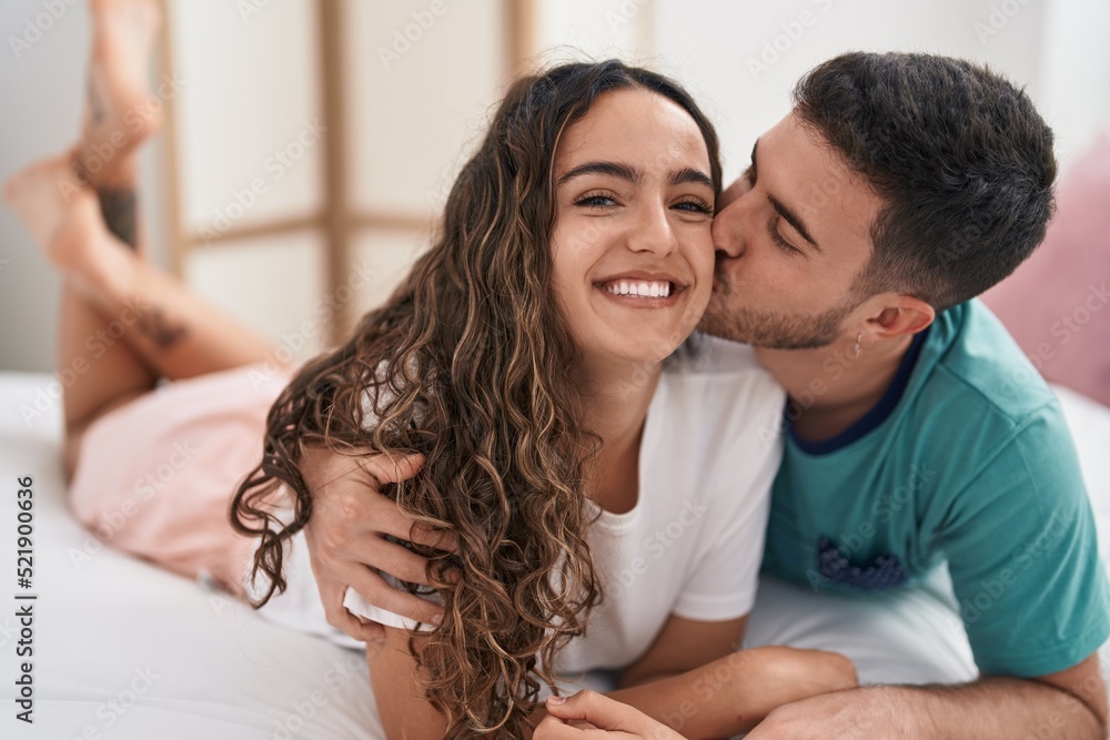 Young hispanic couple lying on bed hugging each other and kissing at bedroom