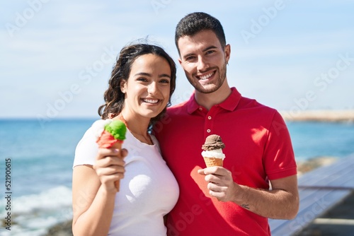 Young hispanic couple tourists hugging each other eating ice cream at seaside