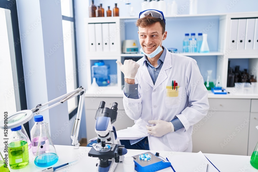 Caucasian man working at scientist laboratory pointing thumb up to the side smiling happy with open mouth Caucasian man working at scientist laboratory pointing thumb up to the side smiling happy with open mouth