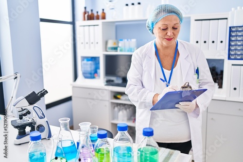 Middle age blonde woman wearing scientist uniform writing on clipboard at laboratory