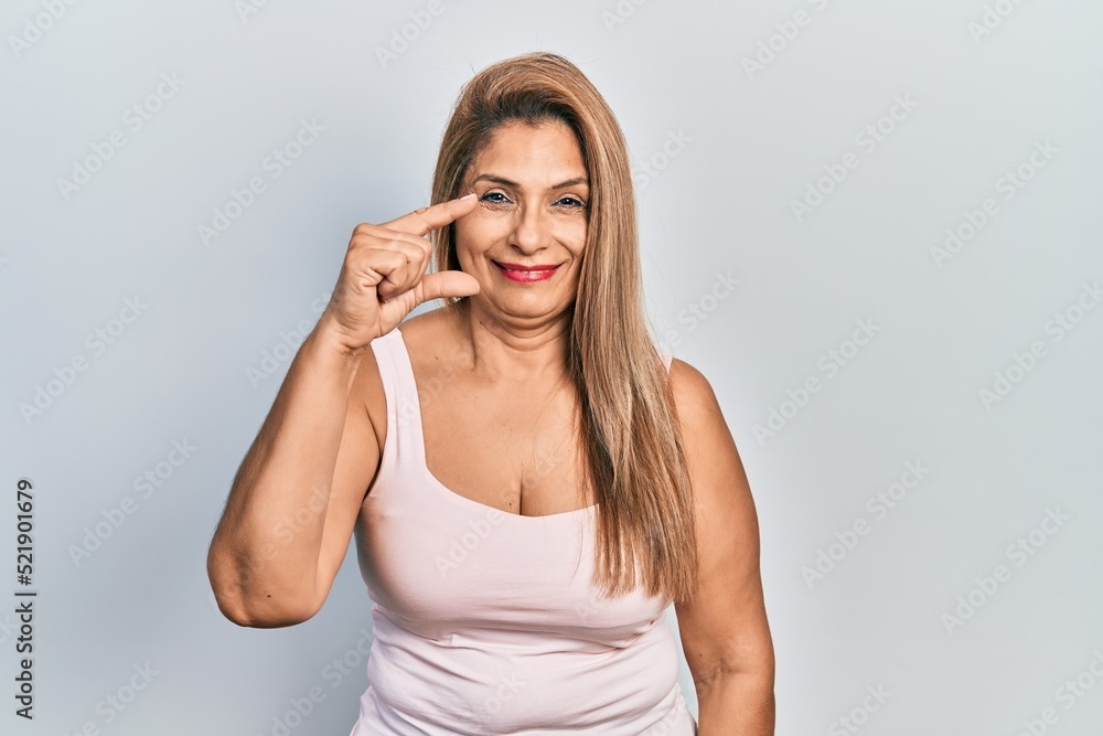 Middle age hispanic woman wearing casual style with sleeveless shirt smiling and confident gesturing with hand doing small size sign with fingers looking and the camera. measure concept. Middle age hispanic woman wearing casual style with sleeveless shirt smiling and confident gesturing with hand doing small size sign with fingers looking and the camera. measure concept.