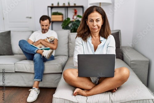 Hispanic middle age couple at home, woman using laptop skeptic and nervous, disapproving expression on face with crossed arms. negative person.