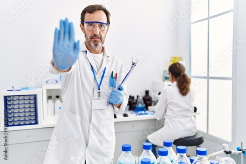 Middle age man working at scientist laboratory with open hand doing stop sign with serious and confident expression  defense gesture