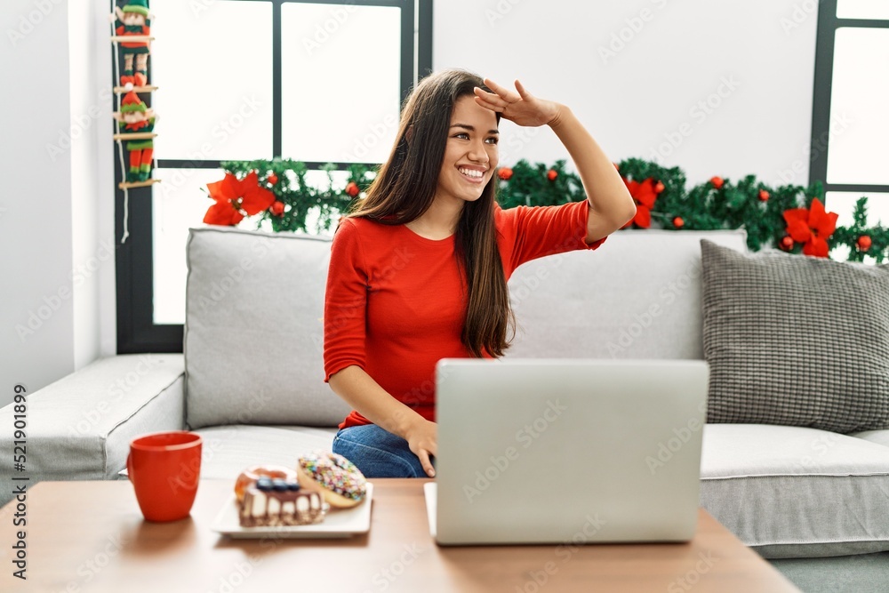 Young brunette woman using laptop sitting on the sofa on christmas very happy and smiling looking far away with hand over head. searching concept. Young brunette woman using laptop sitting on the sofa on christmas very happy and smiling looking far away with hand over head. searching concept.
