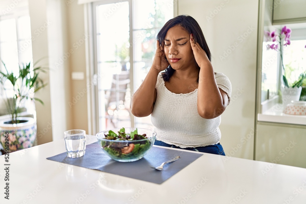 Young hispanic woman eating healthy salad at home with hand on head for pain in head because stress. suffering migraine.