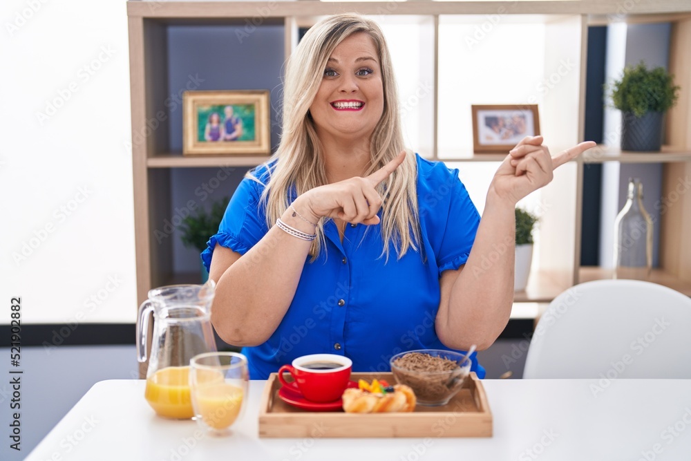 Caucasian plus size woman eating breakfast at home smiling and looking at the camera pointing with two hands and fingers to the side.