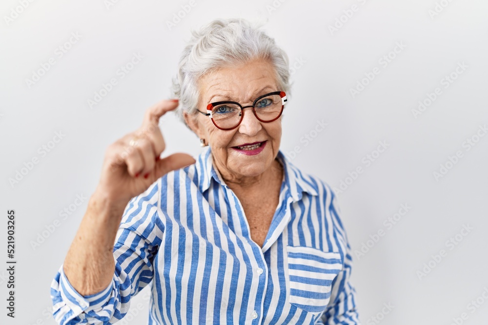 Senior woman with grey hair standing over white background smiling and confident gesturing with hand doing small size sign with fingers looking and the camera. measure concept. Senior woman with grey hair standing over white background smiling and confident gesturing with hand doing small size sign with fingers looking and the camera. measure concept.