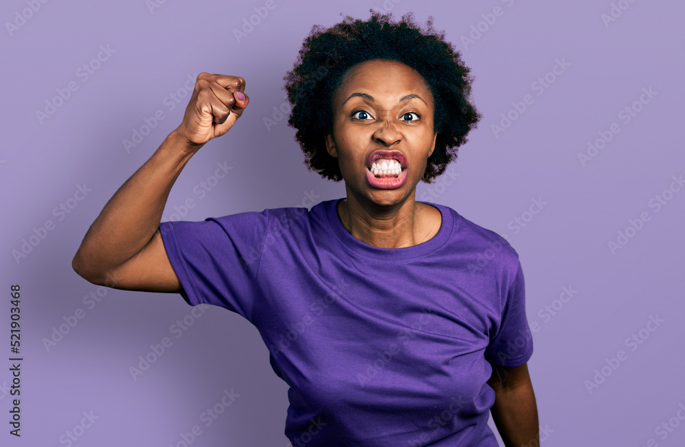 African american woman with afro hair wearing casual purple t shirt angry and mad raising fist frustrated and furious while shouting with anger. rage and aggressive concept. - obrazy, fototapety, plakaty 