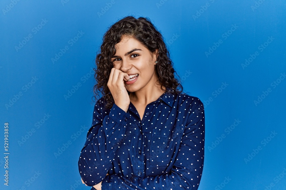 Young brunette woman with curly hair wearing casual clothes over blue background looking stressed and nervous with hands on mouth biting nails. anxiety problem. Young brunette woman with curly hair wearing casual clothes over blue background looking stressed and nervous with hands on mouth biting nails. anxiety problem.