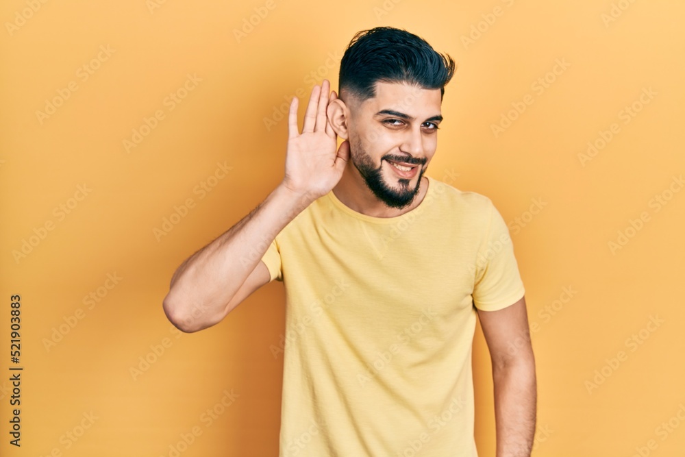 Handsome man with beard wearing casual yellow t shirt smiling with hand over ear listening an hearing to rumor or gossip. deafness concept. Handsome man with beard wearing casual yellow t shirt smiling with hand over ear listening an hearing to rumor or gossip. deafness concept.