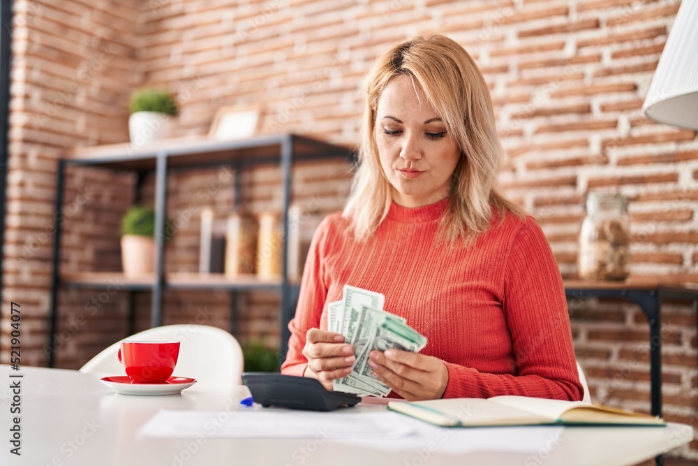 Young blonde woman sitting on table counting dollars at home Young blonde woman sitting on table counting dollars at home