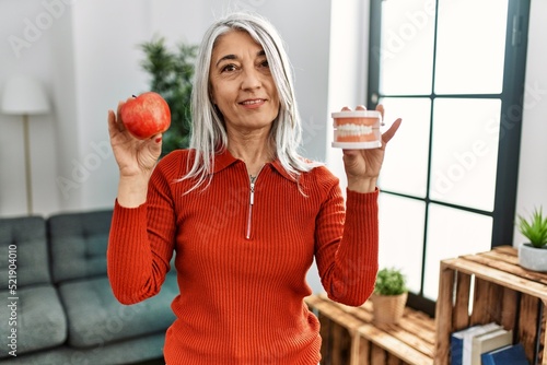 Middle age grey-haired woman smiling confident holding red apple and denture at home photo