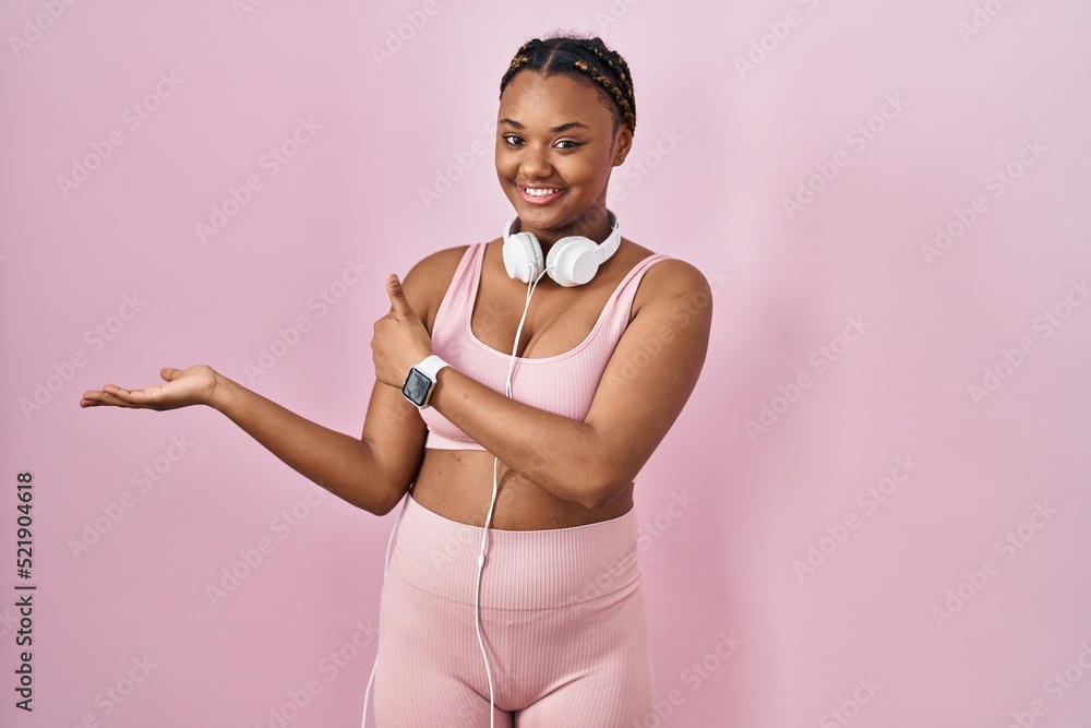 African american woman with braids wearing sportswear and headphones showing palm hand and doing ok gesture with thumbs up, smiling happy and cheerful