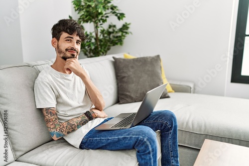 Hispanic man with beard sitting on the sofa looking confident at the camera smiling with crossed arms and hand raised on chin. thinking positive.