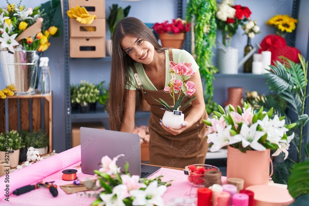 Young beautiful hispanic woman florist using laptop holding plant at flower shop Young beautiful hispanic woman florist using laptop holding plant at flower shop