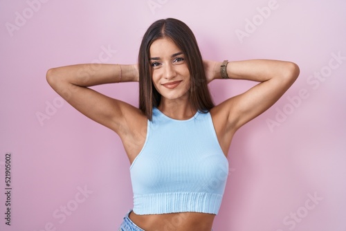 Young brunette woman standing over pink background relaxing and stretching, arms and hands behind head and neck smiling happy © Krakenimages.com