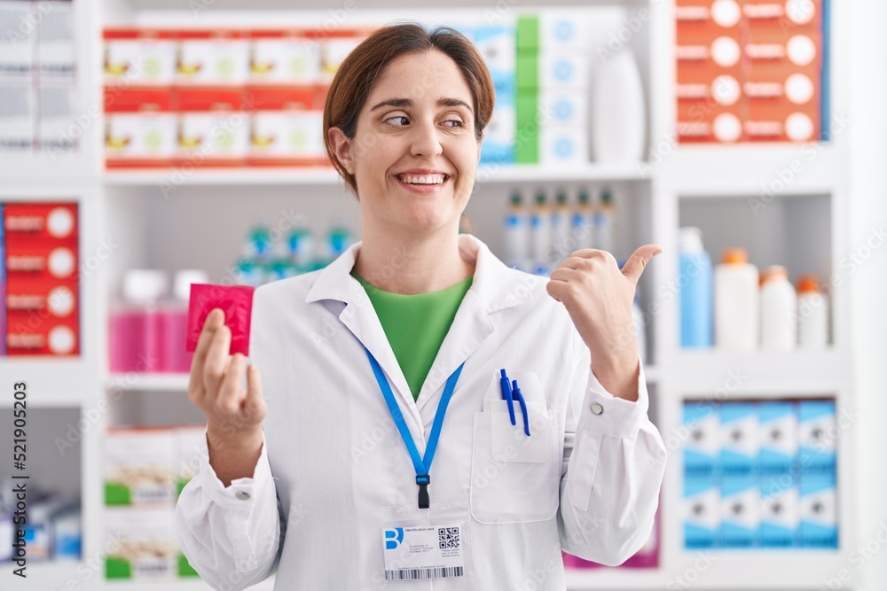 Brunette woman working at pharmacy drugstore holding condom pointing thumb up to the side smiling happy with open mouth Brunette woman working at pharmacy drugstore holding condom pointing thumb up to the side smiling happy with open mouth