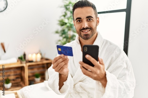 Young hispanic man wearing bathrobe using smartphone and credit card at beauty center