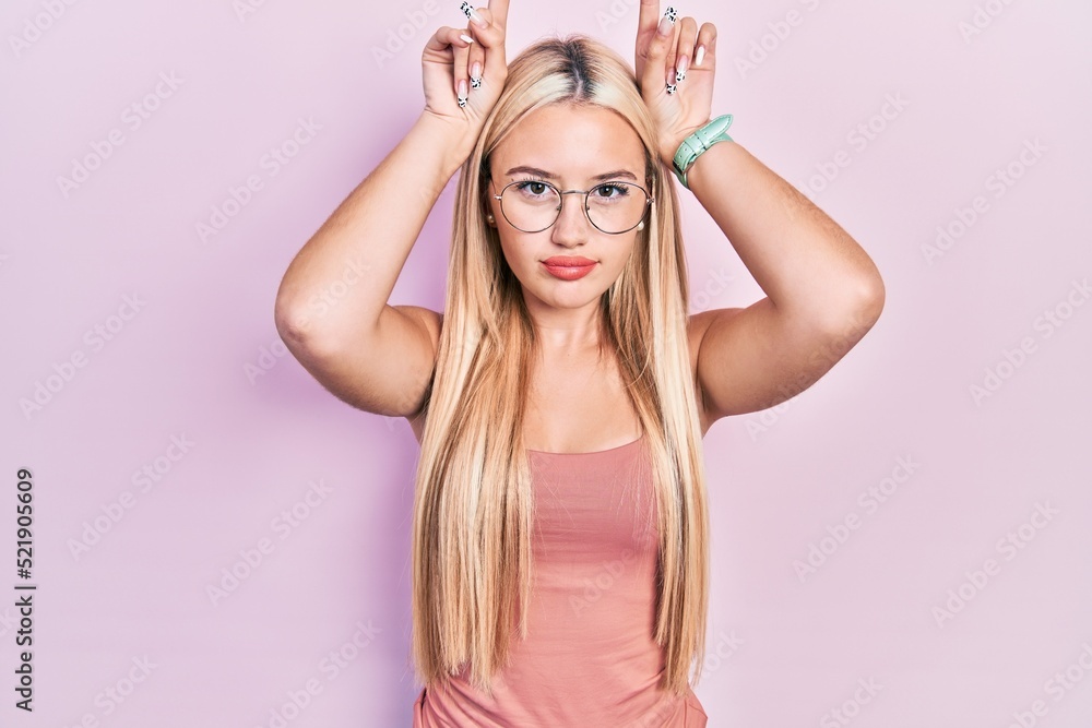 Young blonde girl wearing casual clothes doing funny gesture with finger over head as bull horns Young blonde girl wearing casual clothes doing funny gesture with finger over head as bull horns