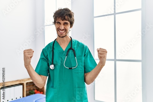 Young hispanic man wearing doctor uniform and stethoscope at clinic very happy and excited doing winner gesture with arms raised, smiling and screaming for success. celebration concept.