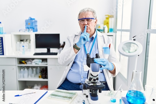 Senior caucasian man working at scientist laboratory asking to be quiet with finger on lips. silence and secret concept.