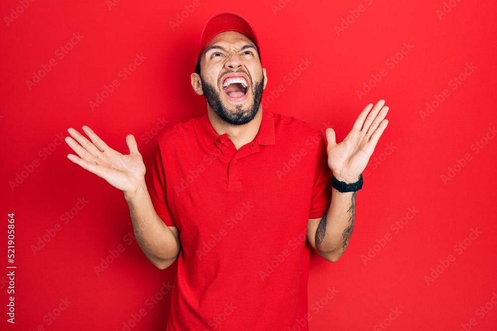 Naklejka premium Hispanic man with beard wearing delivery uniform and cap celebrating mad and crazy for success with arms raised and closed eyes screaming excited. winner concept