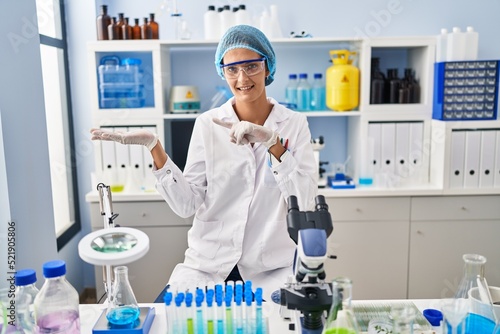 Brunette woman working at scientist laboratory amazed and smiling to the camera while presenting with hand and pointing with finger.