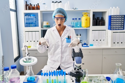 Brunette woman working at scientist laboratory pointing down with fingers showing advertisement, surprised face and open mouth