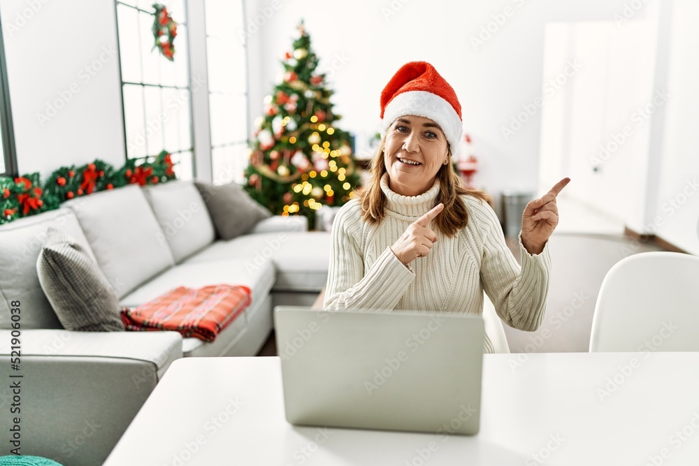 Middle age woman wearing santa claus hat using laptop smiling and looking at the camera pointing with two hands and fingers to the side. Middle age woman wearing santa claus hat using laptop smiling and looking at the camera pointing with two hands and fingers to the side.