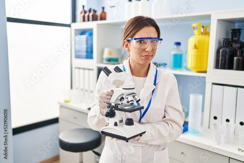 Young brunette woman working at scientist laboratory with microscope skeptic and nervous, frowning upset because of problem. negative person.