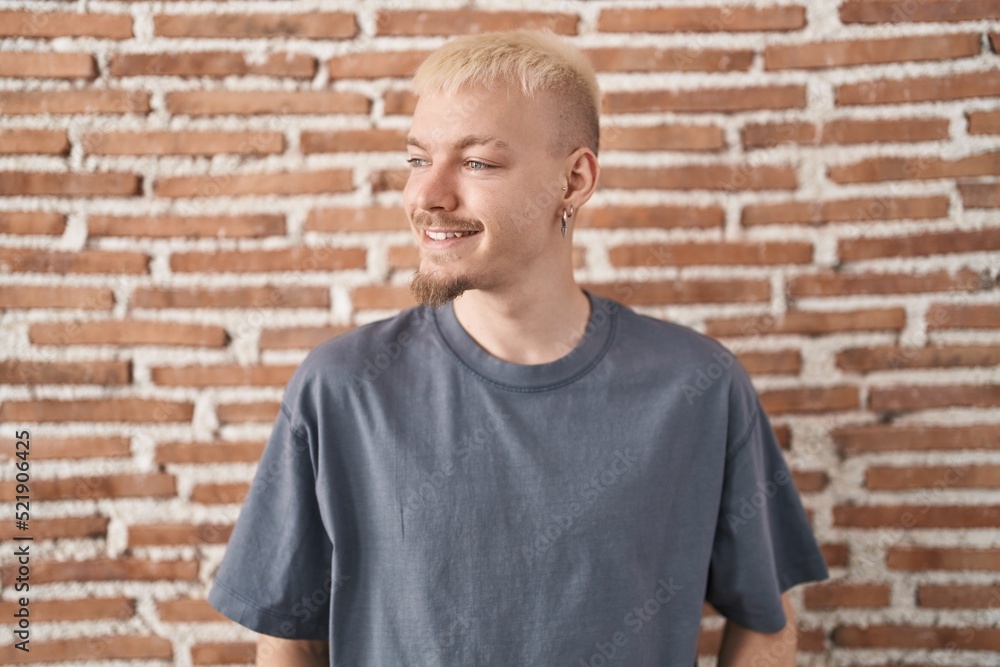 Young caucasian man standing over bricks wall looking away to side with smile on face, natural expression. laughing confident. Young caucasian man standing over bricks wall looking away to side with smile on face, natural expression. laughing confident.