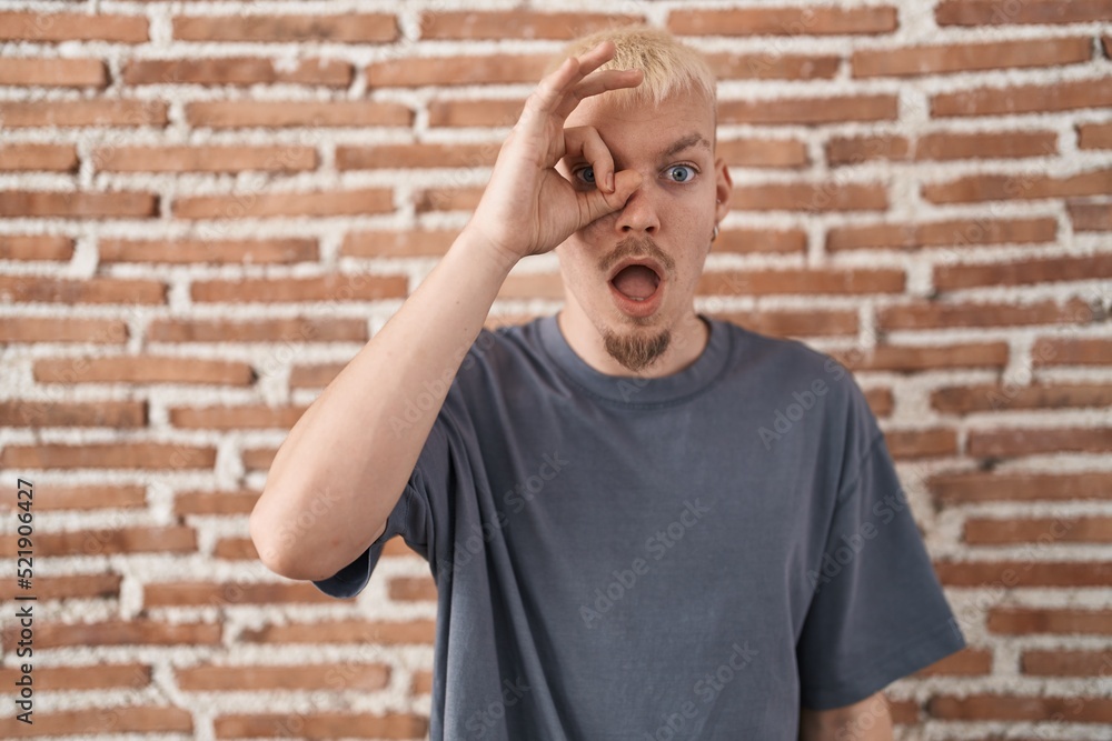 Young caucasian man standing over bricks wall doing ok gesture shocked with surprised face, eye looking through fingers. unbelieving expression. Young caucasian man standing over bricks wall doing ok gesture shocked with surprised face, eye looking through fingers. unbelieving expression.