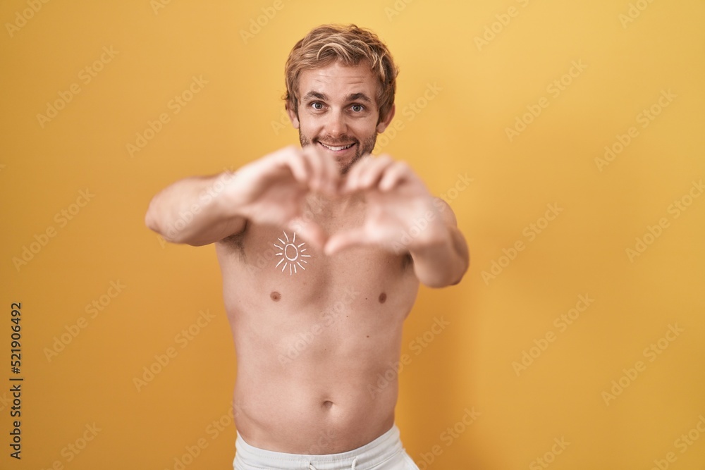 Caucasian man standing shirtless wearing sun screen smiling in love doing heart symbol shape with hands. romantic concept.