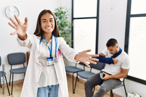 Young asian doctor woman at waiting room with a man with a broken arm looking at the camera smiling with open arms for hug. cheerful expression embracing happiness.