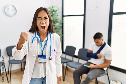 Young asian doctor woman at waiting room with a man with a broken arm angry and mad raising fist frustrated and furious while shouting with anger. rage and aggressive concept.