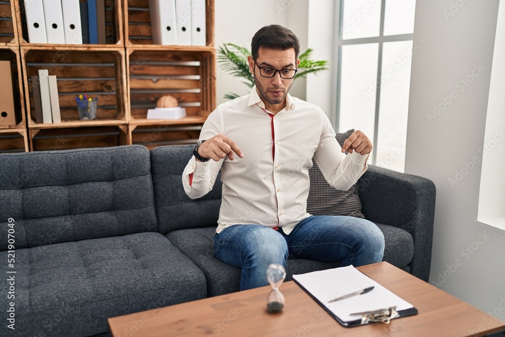 Young hispanic man with beard working at consultation office pointing down with fingers showing advertisement, surprised face and open mouth Young hispanic man with beard working at consultation office pointing down with fingers showing advertisement, surprised face and open mouth