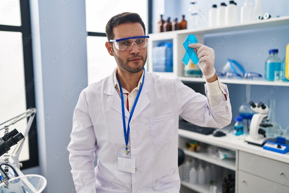 Young hispanic man with beard working at scientist laboratory holding blue ribbon thinking attitude and sober expression looking self confident Young hispanic man with beard working at scientist laboratory holding blue ribbon thinking attitude and sober expression looking self confident