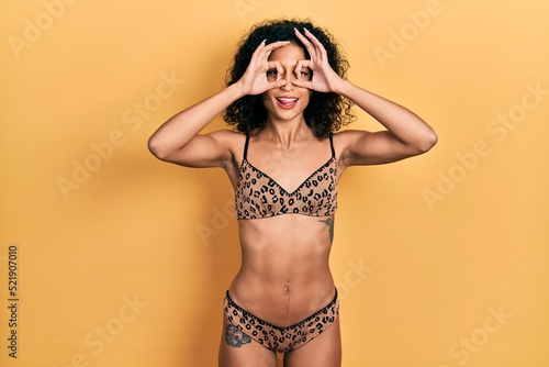 Young latin girl wearing bikini doing ok gesture like binoculars sticking tongue out, eyes looking through fingers. crazy expression.