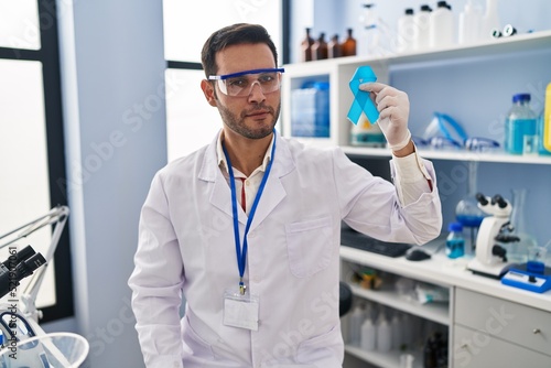 Young hispanic man with beard working at scientist laboratory holding blue ribbon thinking attitude and sober expression looking self confident