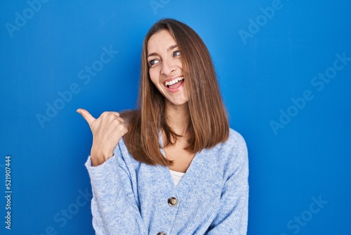 Young woman standing over blue background smiling with happy face looking and pointing to the side with thumb up.