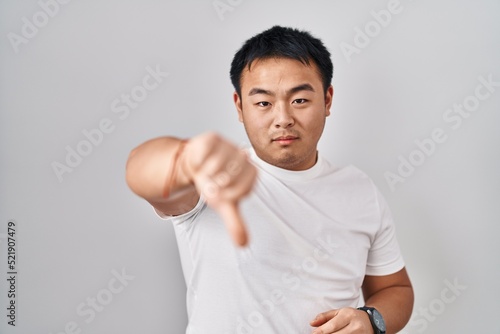 Young chinese man standing over white background looking unhappy and angry showing rejection and negative with thumbs down gesture. bad expression. © Krakenimages.com