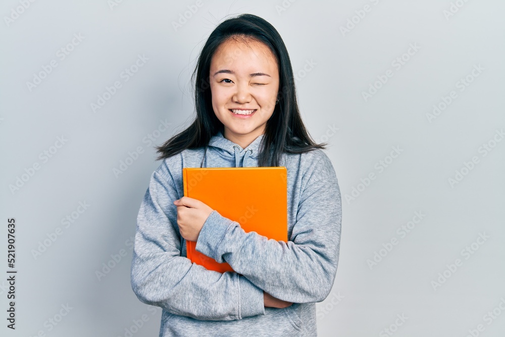 Young chinese girl holding book winking looking at the camera with sexy expression, cheerful and happy face. Young chinese girl holding book winking looking at the camera with sexy expression, cheerful and happy face.